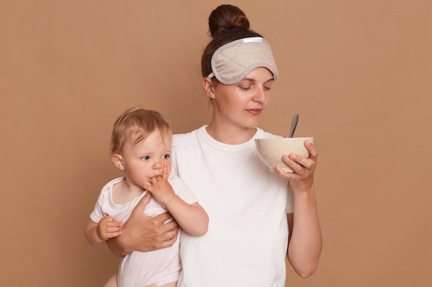 Portrait of woman wearing white t shirt and sleeping mask holding infant kid in hands smelling food in plate for feeding baby posing isolated over brown background