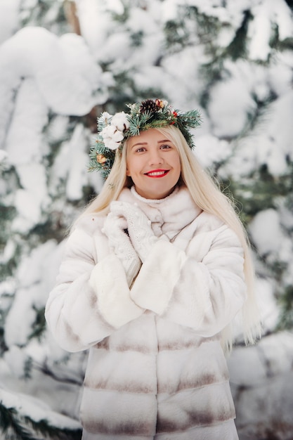 Portrait of a woman in a white fur coat in a cold winter forest. Girl with a wreath on her head in a snow-covered winter forest.