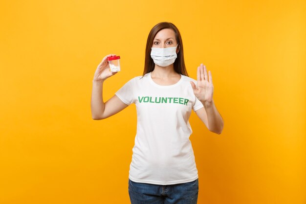 Portrait woman in white t-shirt written inscription green title volunteer, sterile face mask with pills drug isolated on yellow background. Voluntary free assistance help, charity grace health concept