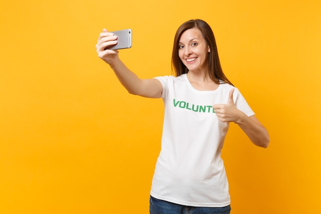 Portrait of woman in white t-shirt written inscription green title volunteer taking selfie shot on mobile phone isolated on yellow background. Voluntary free assistance help charity grace work concept