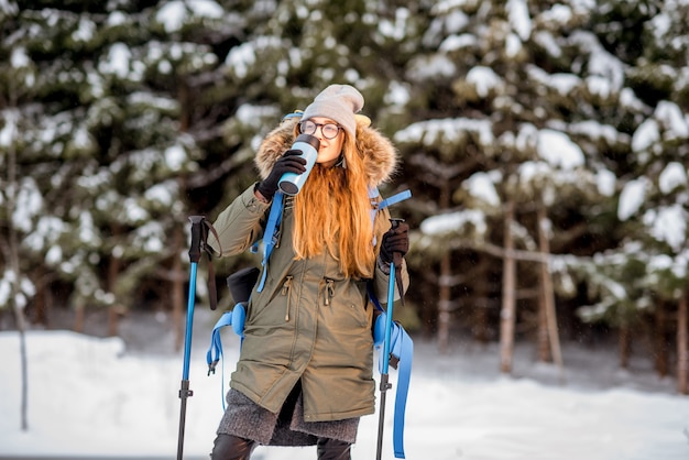 Portrait of a woman in winter clothes hiking with backpack, tracking sticks and thermos at the snowy forest near the frozen lake