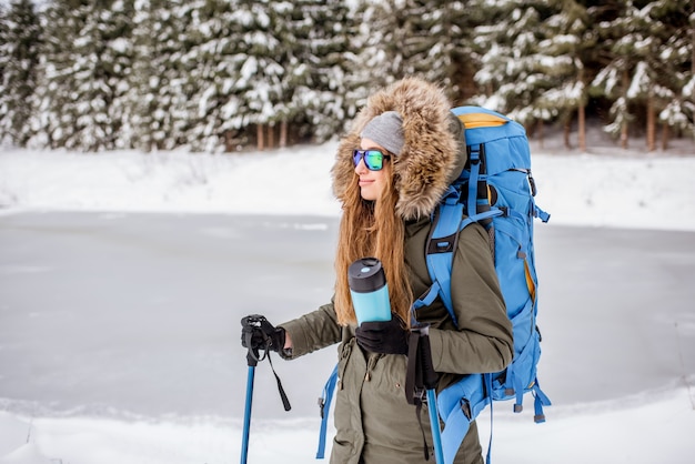 Portrait of a woman in winter clothes hiking with backpack, tracking sticks and thermos at the snowy forest near the frozen lake
