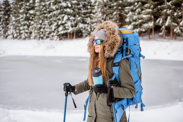 Ritratto di una donna in abiti invernali che fa un'escursione con zaino, bastoncini e thermos nella foresta innevata vicino al lago ghiacciato