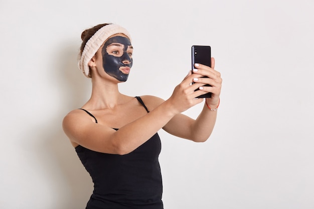 Portrait of woman with black cosmetic mask on her face holding modern smart phone in hand and looking at device screen