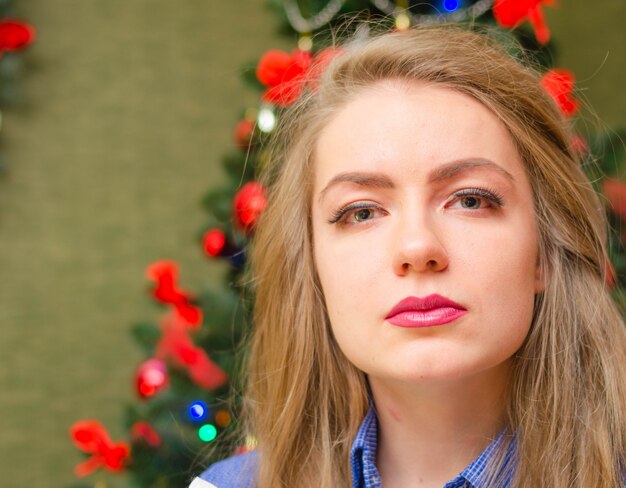 Ritratto di una donna con labbra rosse luminose, capelli lunghi biondi contro l'albero di capodanno. giovane donna in una camicia da uomo blu. vacanze. buon natale. faccia divertente