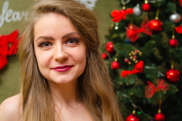 Portrait of a woman with bright red lips, blond long hair against the New Year tree. Young woman in a white warm coat, shoulders are visible. Holidays. Merry Christmas