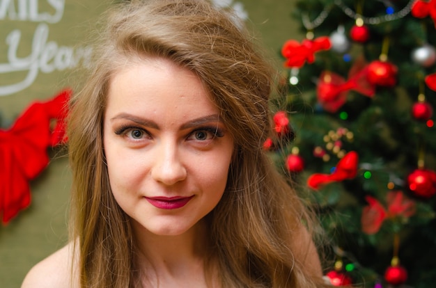 Ritratto di una donna con labbra rosse luminose, capelli lunghi biondi contro l'albero di capodanno. giovane donna in un cappotto bianco caldo, le spalle sono visibili. vacanze. buon natale