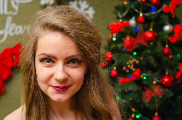 Portrait of a woman with bright red lips, blond long hair against the New Year tree. Young woman in a white warm coat, shoulders are visible. Holidays. Merry Christmas