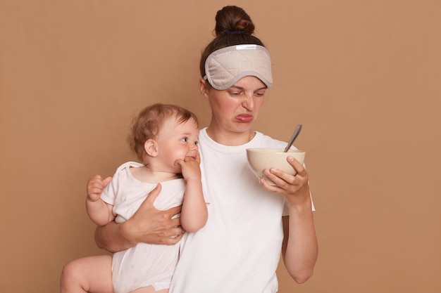 Portrait of woman with disgusted facial expression smelling food in plate preparing dinner for her infant baby daughter mother wearing white t shirt posing with kid isolated over brown background