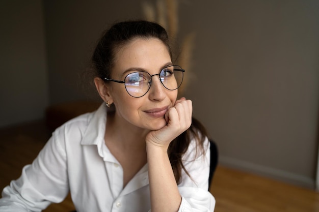 Портрет женщины в очках, смотрящей в камеру в белой рубашке, сидящей в офисе