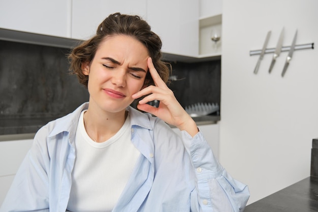 Portrait of woman with headache sits in kitchen touches head and grimaces has painful migraine