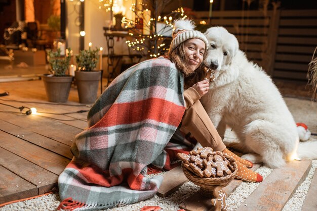 Portrait of a woman with her cute dog celebrating a New Year holidays, sitting together and eating gingerbreads on the beautifully decorated terrace at home