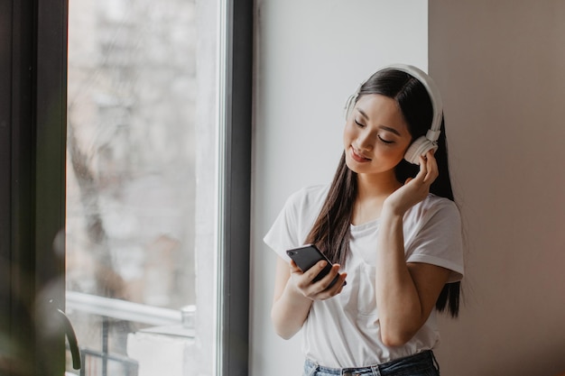 Portrait of woman with long dark hair near window Girl in white tshirt and headphones holds smartphone