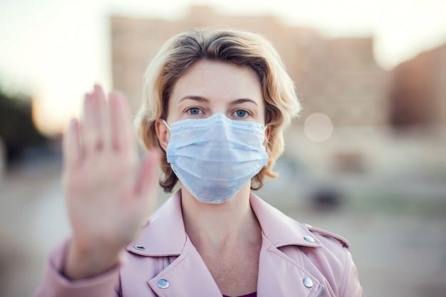 A portrait of woman with medical face mask showing stop sign with hands outdoor. People, healthcare and medicine concept