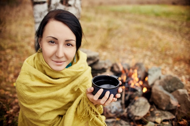 Portrait of a woman with a mug of hot tea in his hands autumn\
in a forest campfire. a picnic in the autumn forest. girl wrapped\
in a blanket warmed in a forest bonfire