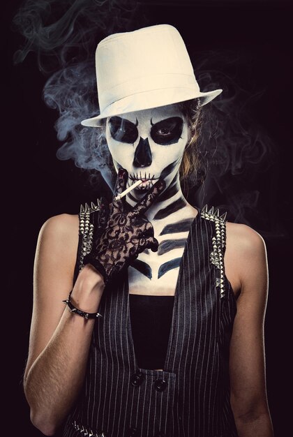 Photo portrait of woman with skull face paint while smoking cigarette against black background