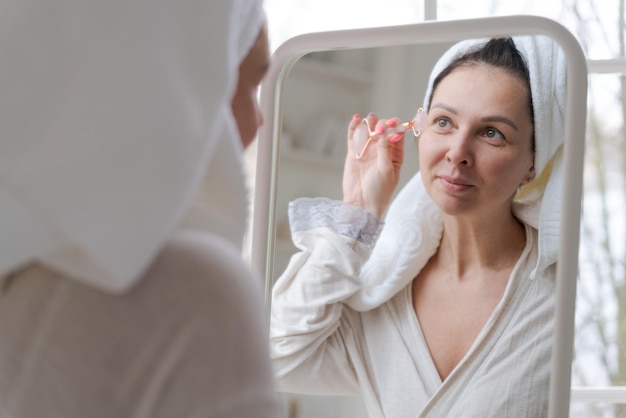 하얀 수건과 목욕 가운에 싸인 초상화 여성은 거울에 비친 거울을 쳐다보며 Y자형 볼 롤러로 얼굴을 마사지합니다. 천연 화장품 개념 주름 완화 선택적 초점