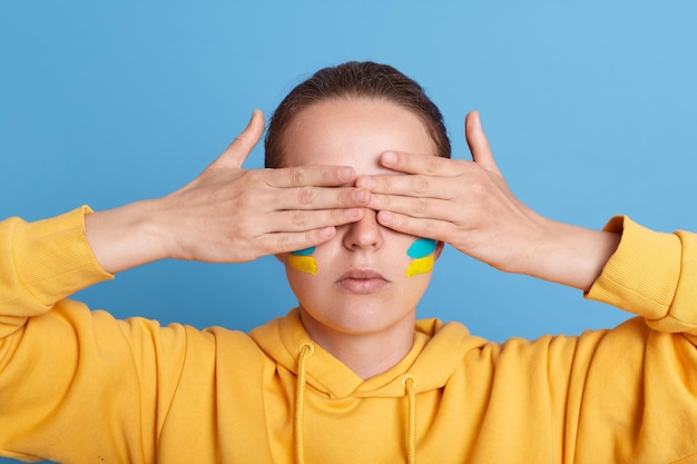Portrait of woman in yellow hoodie with Ukrainian flag on cheeks posing isolated over blue background covering her eyes with palms does not want to see what is happening in her homeland