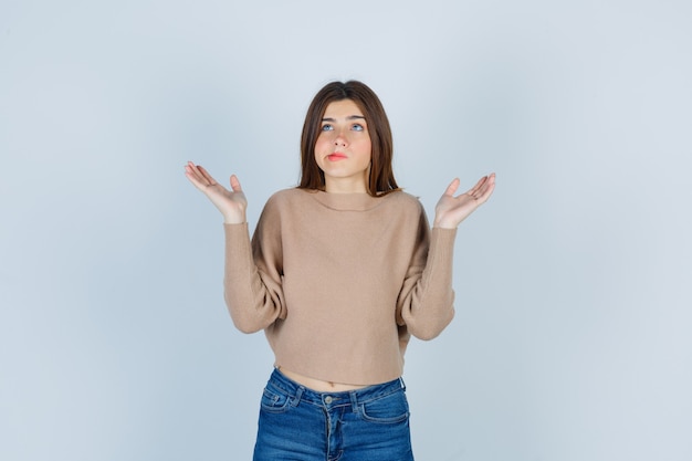 Ritratto di una donna meravigliosa che mostra un gesto impotente, alzando lo sguardo in maglione, jeans e guardando sconcertato la vista frontale