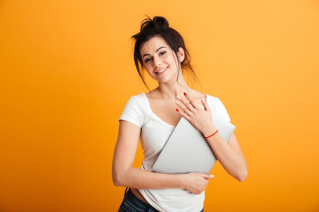 Foto ritratto della donna meravigliosa con l'acconciatura d'avanguardia che abbraccia il computer d'argento moderno e che osserva sulla macchina fotografica con il sorriso schietto, sopra lo spazio giallo della copia della parete