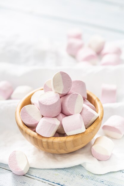 Photo portrait of a wooden bowl full of pink and white marshmallows with some scattered around on a white table cloth.