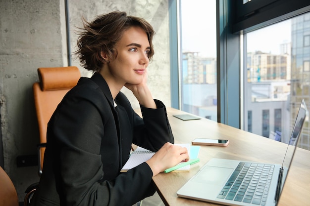 Portrait of working woman in an office using laptop and mobile phone sitting near the window in