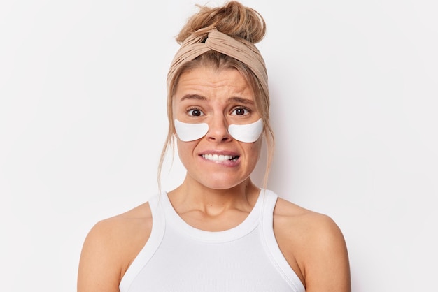 Portrait of worried anxious woman bites lips looks embarrassed feels nervous applies beauty patches under eye to remove dark circles wears headband and t shirt isolated over white background