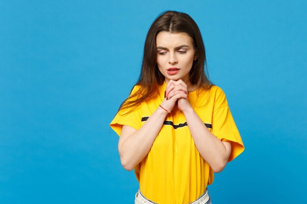 Portrait of worried young woman in vivid casual clothes looking down, holding hands crossed, praying isolated on bright blue wall background in studio. People lifestyle concept. Mock up copy space.