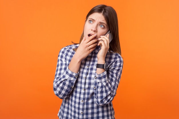 Portrait of worried young woman with brown hair wearing checkered shirt standing talking on smartphone looking up and having pleasant conversation indoor studio shot isolated on orange background