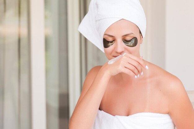 Portrait of wrapped in towel woman with eye patches is applying cream on her hands sitting at the table at the hotel terrace Girl is enjoying a smell Beauty treatment concept