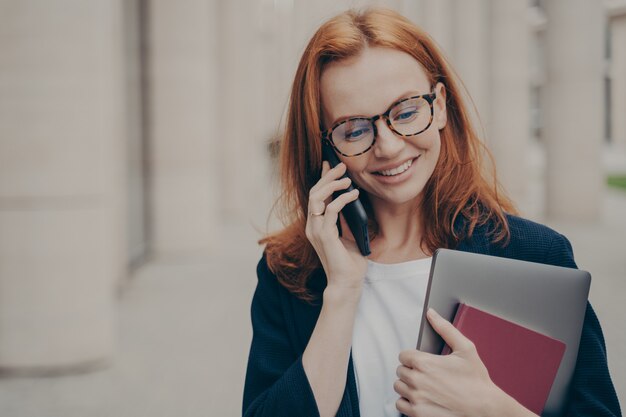 Portrait of young 30s caucasian business woman standing outdoors and talking on smartphone with client, arranging meeting with partner on mobile phone with smile on face, blurred city background