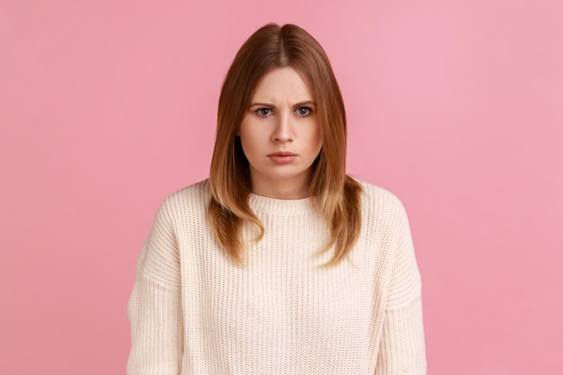 Portrait of young adult angry serious blond woman looking at camera with strict negative expression, aggression, wearing white sweater. Indoor studio shot isolated on pink background.