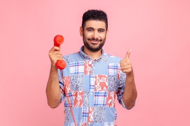 Portrait of young adult attractive man with dark hair and beard showing thumb up and holding red landline phone good service in call center Indoor studio shot isolated on pink background