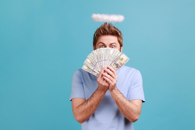 Portrait of young adult bearded angelic man with holy nimbus over head covering half of face with fan of dollar bills, happy to have money. Indoor studio shot isolated on blue background.