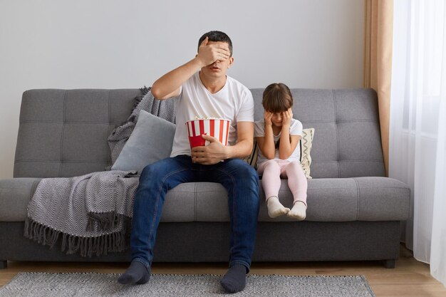 Portrait of young adult Caucasian man watching horror film at home with his child family covering eyes while sees scared moments sitting on sofa and holding popcorn in bucket