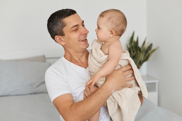 Ritratto di giovane padre adulto con la sua bambina avvolta in un asciugamano, giocando insieme a casa, bell'uomo che guarda il suo bambino neonato con un sorriso a trentadue denti. Foto Premium