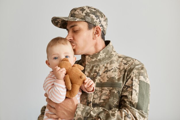 Portrait of young adult military father came from the war or\
the exercises and kissing his toddler daughter, baby holding soft\
toy, male wearing uniform and cap,