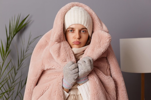 Portrait of young adult sad frozen caucasian woman wearing cap and coat posing in living room trying to get warm in house without central heating wrapped in fur coat