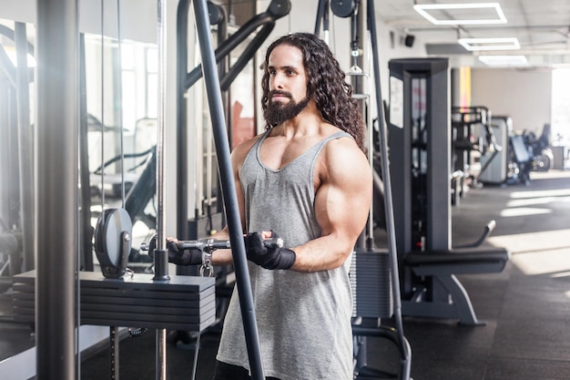 Portrait of young adult sport athlete man with long curly hair training at gym alone standing and lifting weights in the gym doing exercises for biceps Healthy lifestyle concept indoor