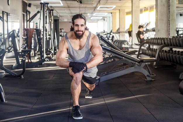 Portrait of young adult sportman athlete with long curly hair working out in gym, squating on one knee, stretching after training, doing exercises for legs, squatting. looking at camera and smiling