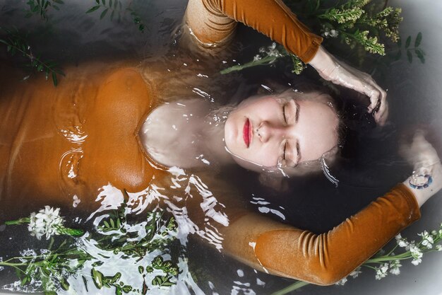 Above portrait of young adult woman lying in bathtub filled with charcoal and flowers