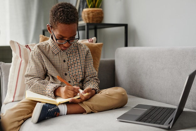 Photo portrait of young african-american boy writing in notebook while studying at home sitting on sofa, copy space