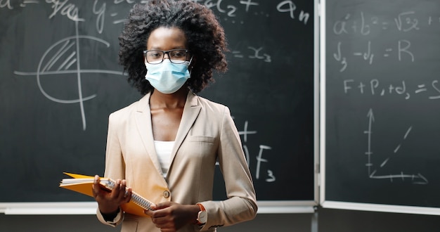 Portrait of young African American female teacher in glasses and medical mask looking at camera in classrom and holding notebooks. Blackboard with formulas on background. Pandemic schooling.