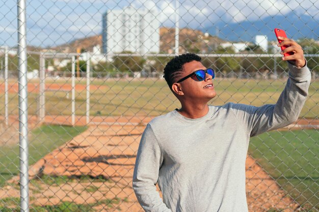 Portrait of a young African American man leaning on a fence, taking a selfie.