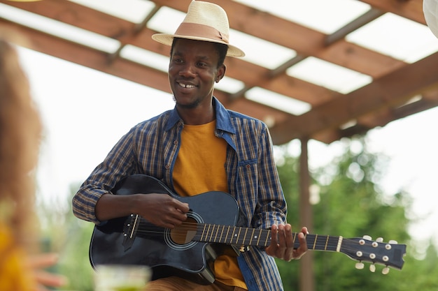 Portrait of young African-American man playing guitar while enjoying dinner with friends outdoors at Summer party