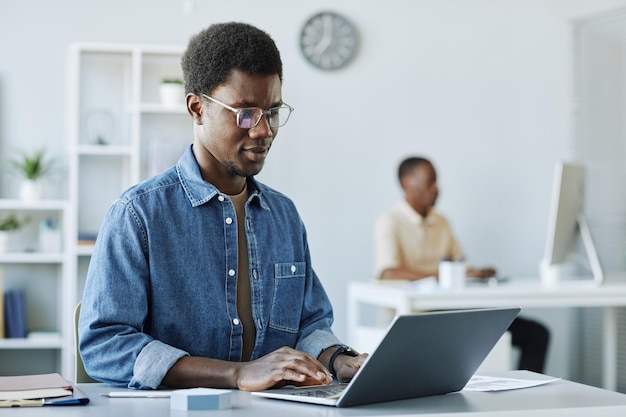 Portrait of young african american man working in office and\
using laptop in minimal grey interior c