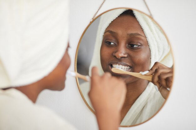 Ritratto di giovane donna afro-americana che si lava i denti al mattino e guarda lo specchio, copia spazio