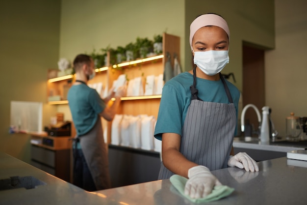 Portrait of young African-American woman cleaning bar counter with sanitizer while working in cafe or coffee shop, covid safety measures, copy space