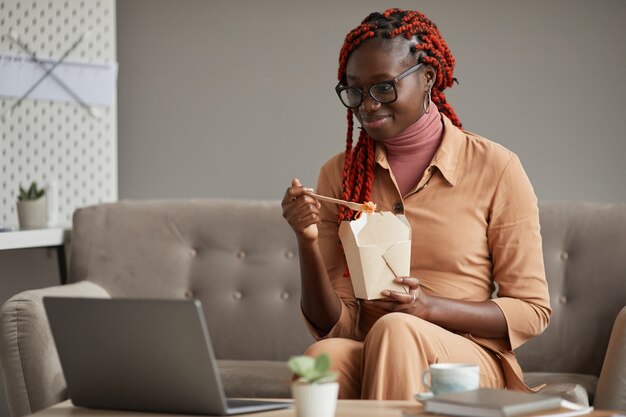 Portrait of young African-American woman eating takeout food and looking at laptop screen while relaxing at home office, copy space