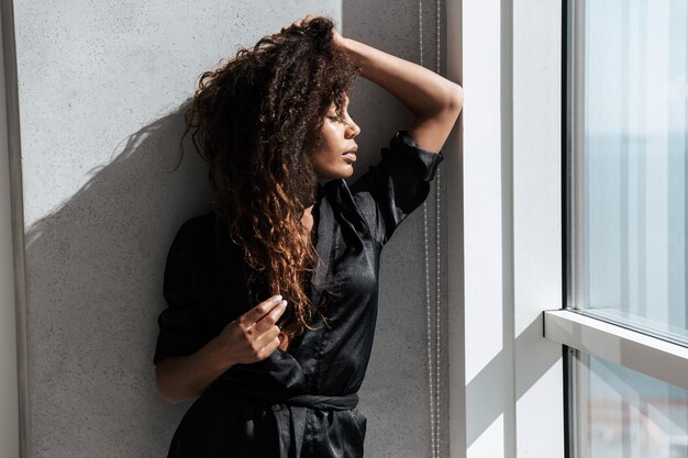 Portrait of young african american woman wearing housecoat, standing near window indoor